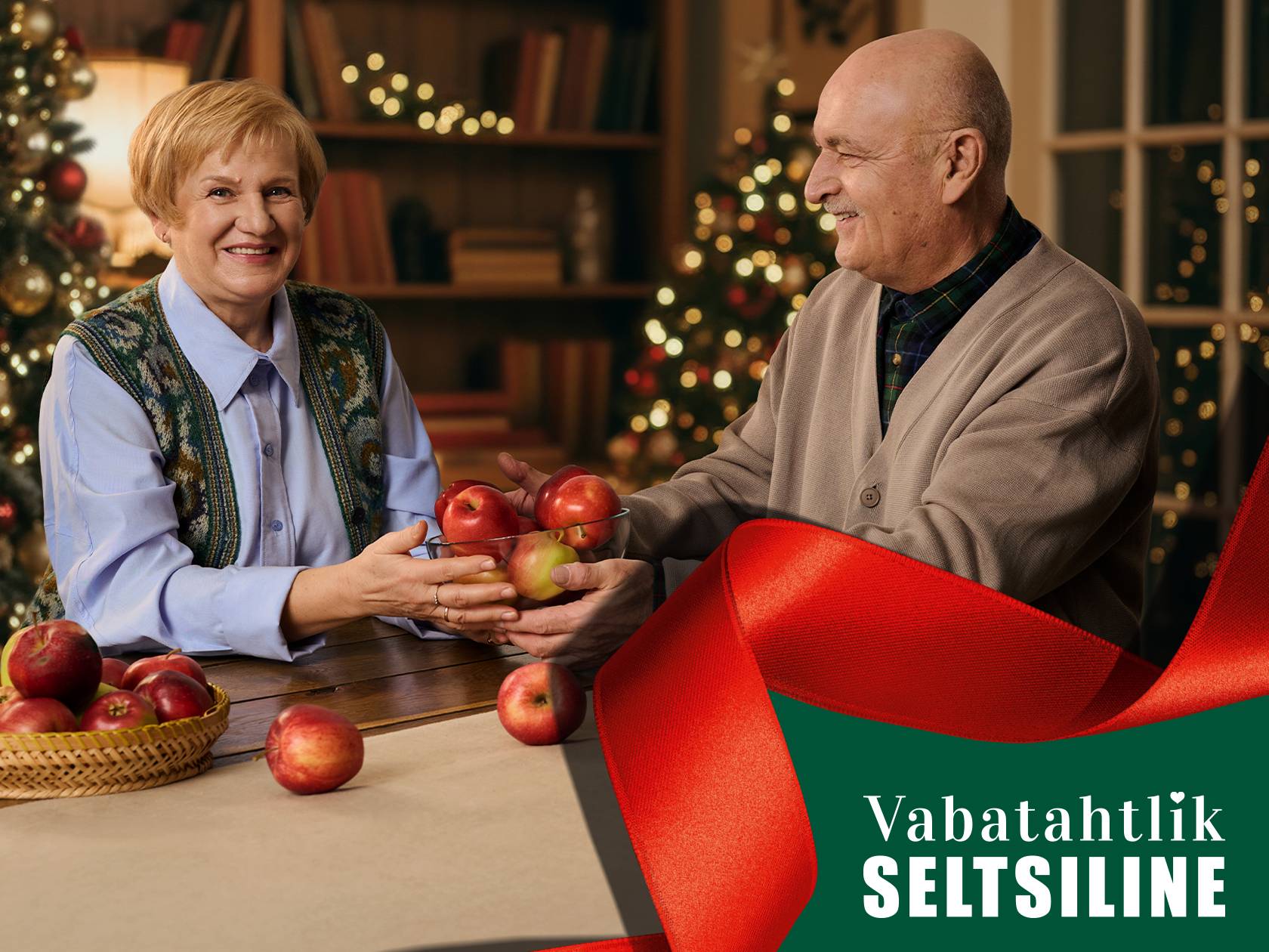 Eakas paar vahetab õunu jõulukaunistuste taustal. Tekst: 'Vabatahtlik SELTSILINE'.