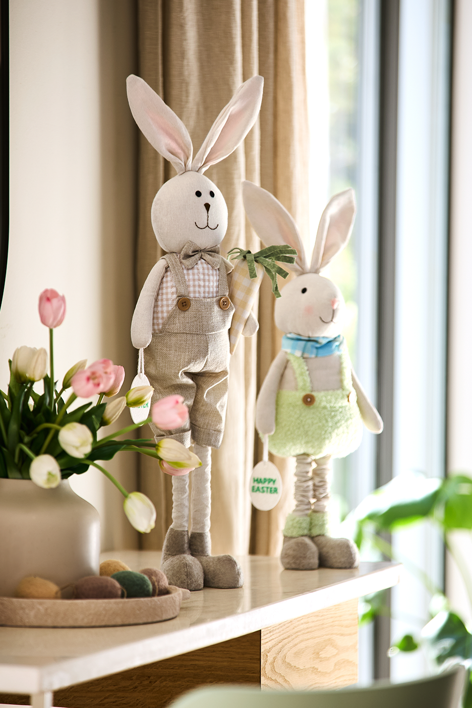 Kaks lihavõttejänest, tulbid ja dekoratiivsed munad laual. Ühel jänesel on silt kirjaga 'Happy Easter'.