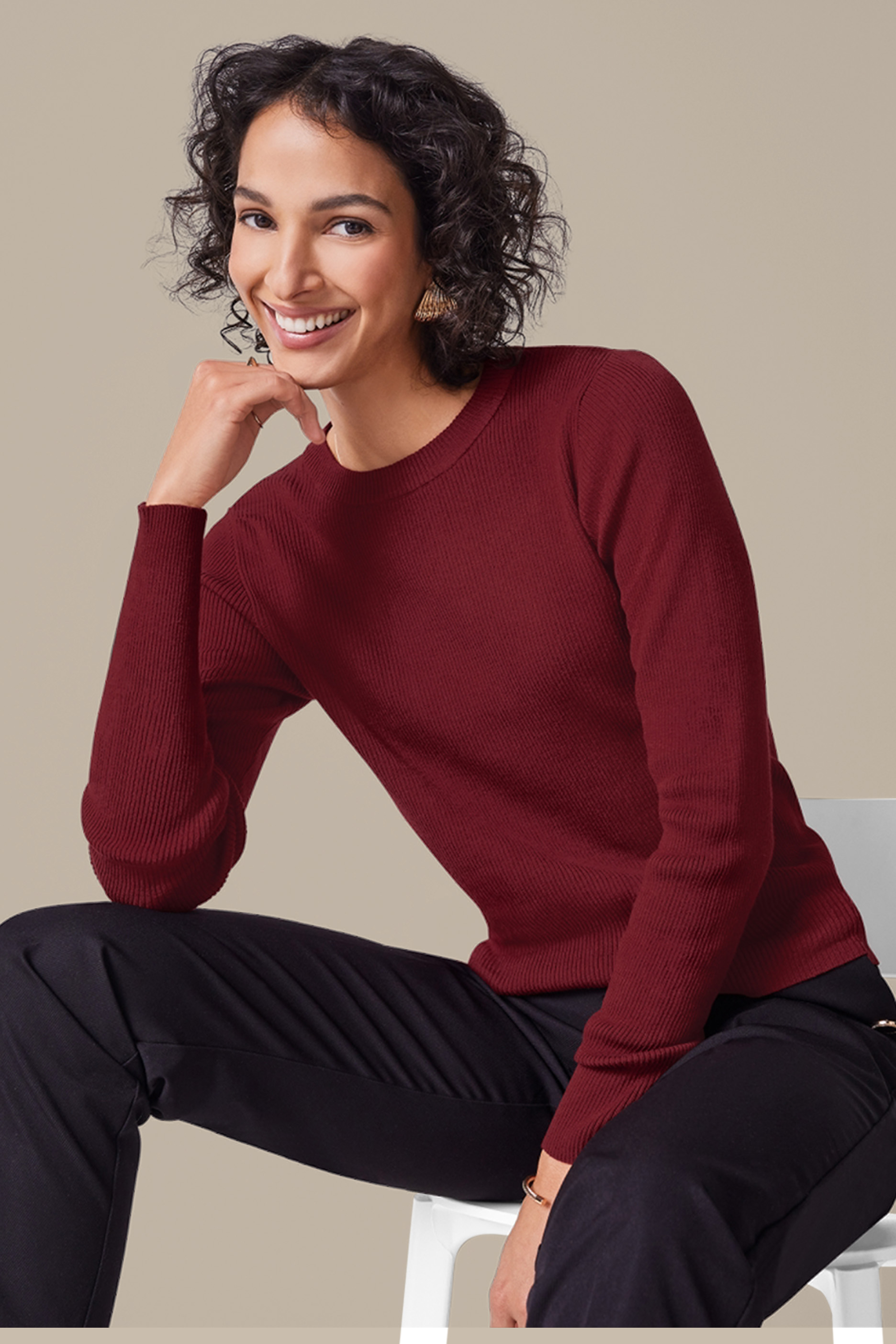 Naine bordoopunases soonilises kampsunis ja mustades pükstes istub toolil ja naeratab.