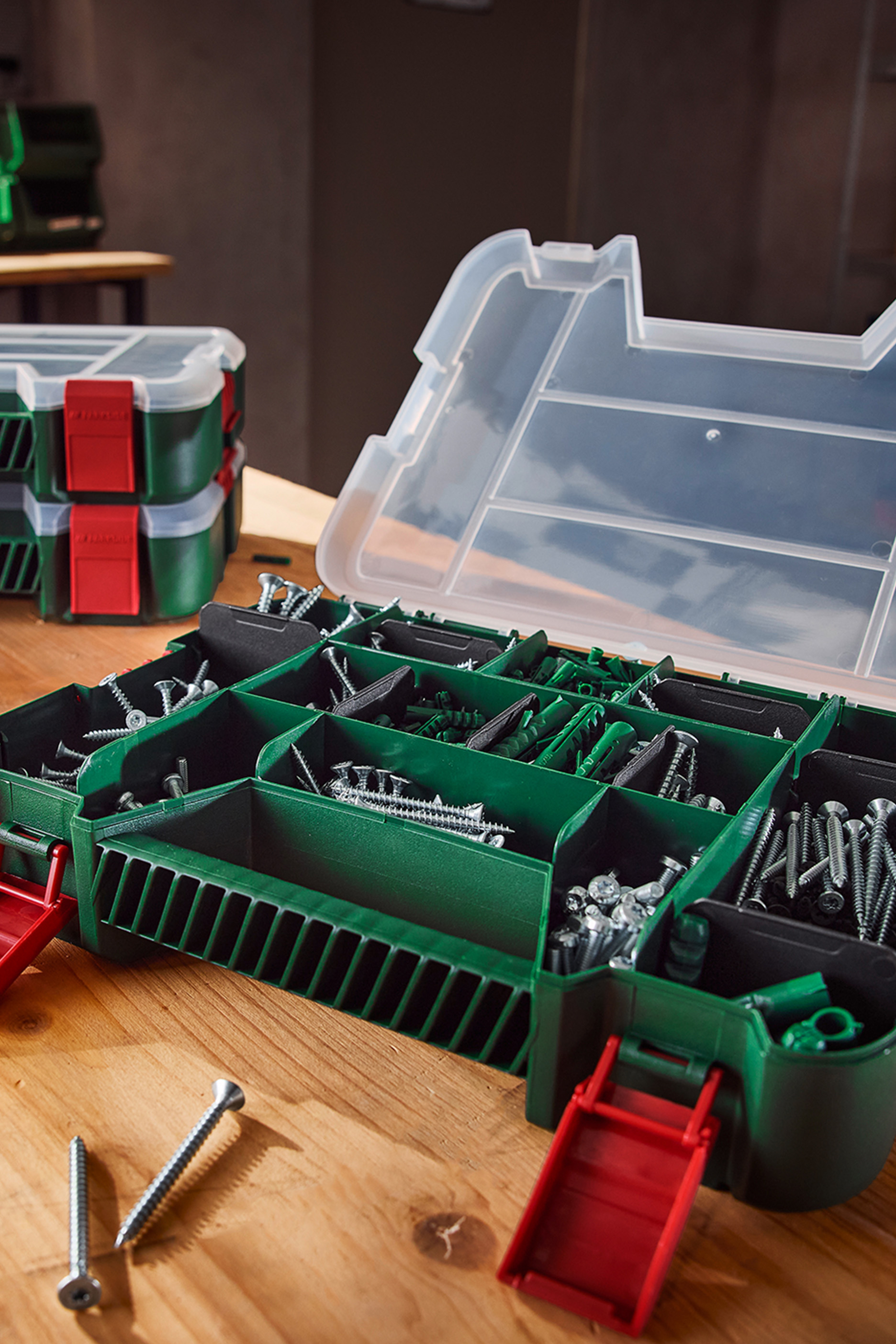 Roheline tööriistakast kruvide ja tüüblitega puidust laual, taustal teised kastid.